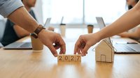 Young adult male and female professionals examining wooden blocks marked 2027 with cardboard house model on desk during business meeting, representing real estate planning, investment strategy, and future property development goals.