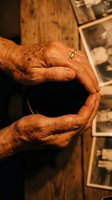 Elderly hands with a gold wedding ring cupped together over a wooden table with vintage black and white photos, evoking memory and life story.