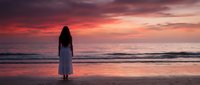 Woman with long dark hair wearing white dress standing alone on beach at sunset, gazing at ocean horizon during golden hour.