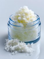 White grainy body scrub in glass jar with small portion on surface, natural skincare product for exfoliation and skin treatment.