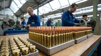 Industrial ammunition manufacturing workers in blue uniforms producing brass cartridges on factory assembly line for defense industry production.