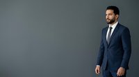 Young Middle Eastern man in formal navy business suit and tie standing against neutral gray background, suitable for corporate, professional, and business-related projects.