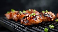 Juicy grilled chicken wings with honey glaze and sesame seeds on black grill grates, garnished with fresh green onions against dark background.