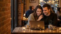 Young couple laughing while using laptop together in cozy cafe with warm lighting and brick walls during evening date.