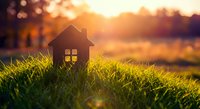 Small wooden house model on green grass at golden sunset representing home ownership dreams and real estate investment opportunities.