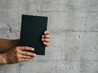 Male hands holding blank black book against concrete wall background, suitable for publishing, education, and marketing mockup designs.