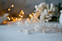 Delicate crystal snowflake hair accessories with pearls on white surface, soft bokeh lights and white flowers in background create magical winter mood.