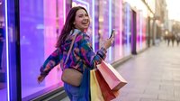 Happy young woman with shopping bags using smartphone while walking past colorful neon-lit storefronts in urban shopping district during evening hours.