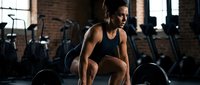 Athletic African American woman in sportswear performing squat exercise in modern gym with brick walls and fitness equipment for strength training workout.
