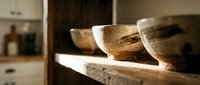 Rustic handmade ceramic bowls on a wooden shelf in a warm, sunlit kitchen interior, showcasing artisan pottery craftsmanship.
