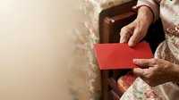 Senior woman with light skin tone holding red envelope, wearing floral patterned clothing, giving gift during celebration or festive occasion, close-up of hands and torso.