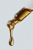 Golden honey or syrup drop falling from glass dropper against white background, capturing viscous liquid movement and light reflection in macro photography.