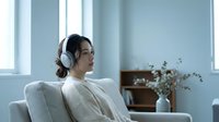 Young Asian woman wearing wireless headphones, sitting on light gray armchair in modern minimalist living room with large windows, dried flowers in vase, listening to music or podcast.