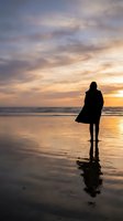 Silhouetted woman standing on wet beach at sunset, gazing at ocean horizon during golden hour. Contemplative mood for wellness, mindfulness, and lifestyle concepts.