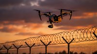 Professional drone flying over security fence with barbed wire during dramatic sunset for surveillance and monitoring operations.