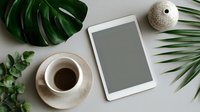 Minimalist workspace with tablet, coffee cup, and tropical leaves on neutral background for modern lifestyle content creation.