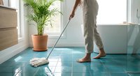 Person mopping bright bathroom floor with natural light streaming through window. Clean modern interior with palm plant and tiled surfaces.