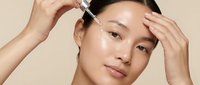 Young Asian woman applying facial serum with dropper for skincare routine and beauty treatment on neutral background.