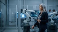 Senior woman with white hair, Caucasian ethnicity, wearing dark business attire, holding tablet in modern industrial facility with robotic arms and manufacturing equipment in background.