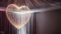 Heart-shaped wire frame pendant light fixture with warm LED bulbs hanging in elegant interior space with draped fabric background for romantic decor.