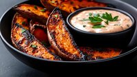 Grilled potato wedges with spicy seasoning and charred edges served in black ceramic plate with creamy dipping sauce garnished with fresh parsley.