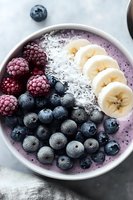 Purple smoothie bowl topped with fresh blueberries, raspberries, sliced banana and shredded coconut on marble background, healthy breakfast concept.