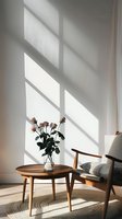 Sunlight casting dramatic shadows on white wall with pink roses in glass vase on wooden side table next to mid-century armchair, creating minimalist interior scene.