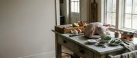 Rustic kitchen counter with fresh vegetables, bread, herbs and cooking ingredients arranged near bright windows in farmhouse style interior.