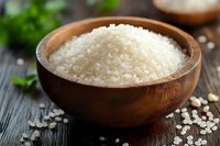 Raw white rice grains in wooden bowl on dark rustic table with herbs and scattered rice. Natural food photography for cooking blogs and culinary websites.