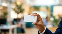 Close-up of male hand holding blank white business card in modern office environment with blurred background, suitable for corporate branding and professional networking.