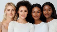 Diverse group of young women with different skin tones and hair types smiling at camera, wearing white tops against neutral background. Natural beauty concept.