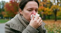 Sick woman blowing nose with tissue during autumn cold and flu season in park with colorful fall foliage background for health and wellness concepts.