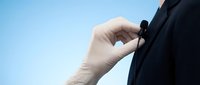 Professional businessman adjusting small microphone on suit lapel against clear blue sky background for corporate communication and presentation.
