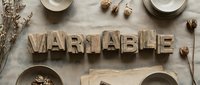 Wooden letter blocks spelling VARIABLE on rustic linen surface with ceramic bowls, dried flowers and natural autumn decor elements for creative projects.