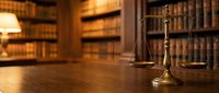 Golden scales of justice on wooden desk in traditional law library with leather-bound legal books creating warm professional atmosphere for legal services.