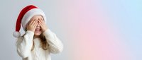 Young woman in Santa hat covering eyes with hands in surprise gesture wearing white sweater on gradient background for holiday marketing campaigns.