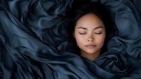 Young Asian woman sleeping peacefully surrounded by flowing dark blue silk fabric creating dreamy atmosphere. Serene facial expression against luxurious textile background.