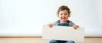 Happy toddler boy with curly hair holding blank white sign while sitting on wooden floor against white wall for advertising and promotional content.