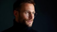 Young Caucasian man with beard wearing round glasses and black turtleneck sweater looking thoughtfully aside against dark background in dramatic lighting.