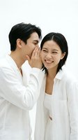 Young Asian couple sharing a secret, man whispering in smiling woman's ear, both in white linen shirts, light background, joyful bond.