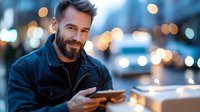 Smiling bearded Caucasian man in denim jacket using digital tablet outdoors at dusk with blurred city lights and bokeh background. Modern lifestyle concept.