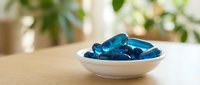 Blue gel capsules supplements in white ceramic bowl on wooden table with green plants background for health and wellness concepts.