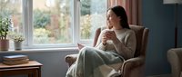 Young Asian woman in cozy sweater relaxing in armchair by window with hot beverage, enjoying peaceful morning moment in comfortable home interior.