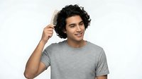Young man with curly dark hair and olive skin tone wearing casual gray t-shirt, touching hair with hand, smiling at camera against white background for lifestyle and fashion content.