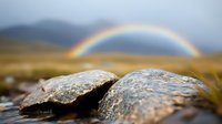 Vibrant rainbow arcs over mountain landscape with weathered granite rocks in foreground, dramatic storm clouds clearing after rain in scenic wilderness.