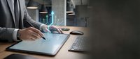 Businessman in suit working on tablet with holographic data analytics chart overlay, modern office workspace with keyboard and laptop visible in background.