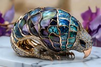 Luxurious brooch shaped as bird with iridescent blue and purple stained glass mosaic wings, adorned with crystals and gold-toned metal details on light background.