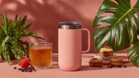 Pink insulated travel mug with handle on coral background surrounded by tropical plants, glass of tea, coffee beans and cookies for morning beverage concept.