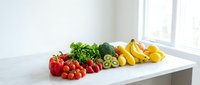 Fresh colorful vegetables and fruits arranged on white kitchen counter near window for healthy cooking and meal preparation.