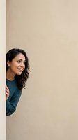 Young woman with long dark wavy hair and South Asian ethnicity peeking around beige wall corner wearing teal sweater, smiling with copy space for advertising.